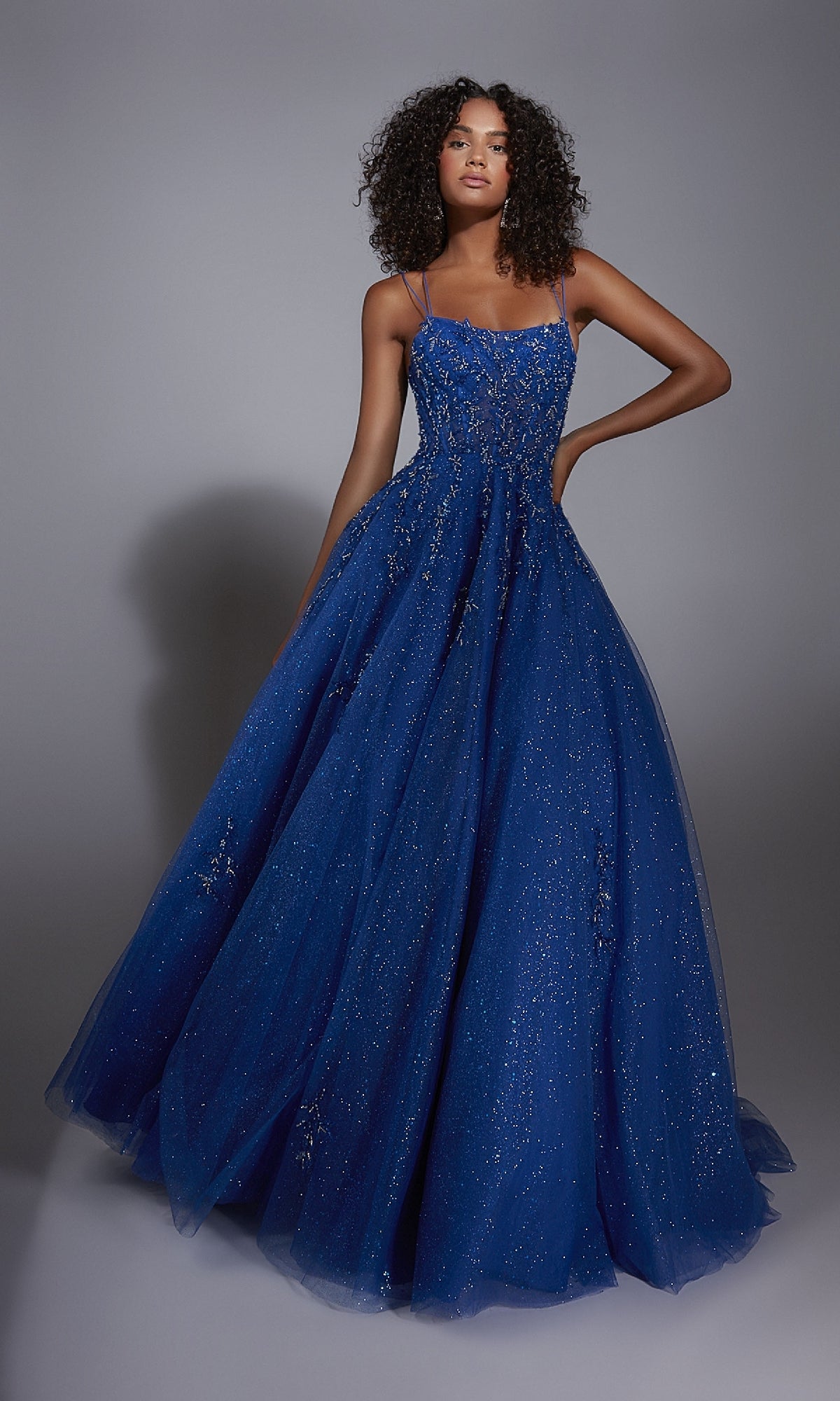 A woman poses against a plain gray background wearing the Alyce 61824, a floor-length blue glitter prom ball gown with thin straps and a corset bodice.